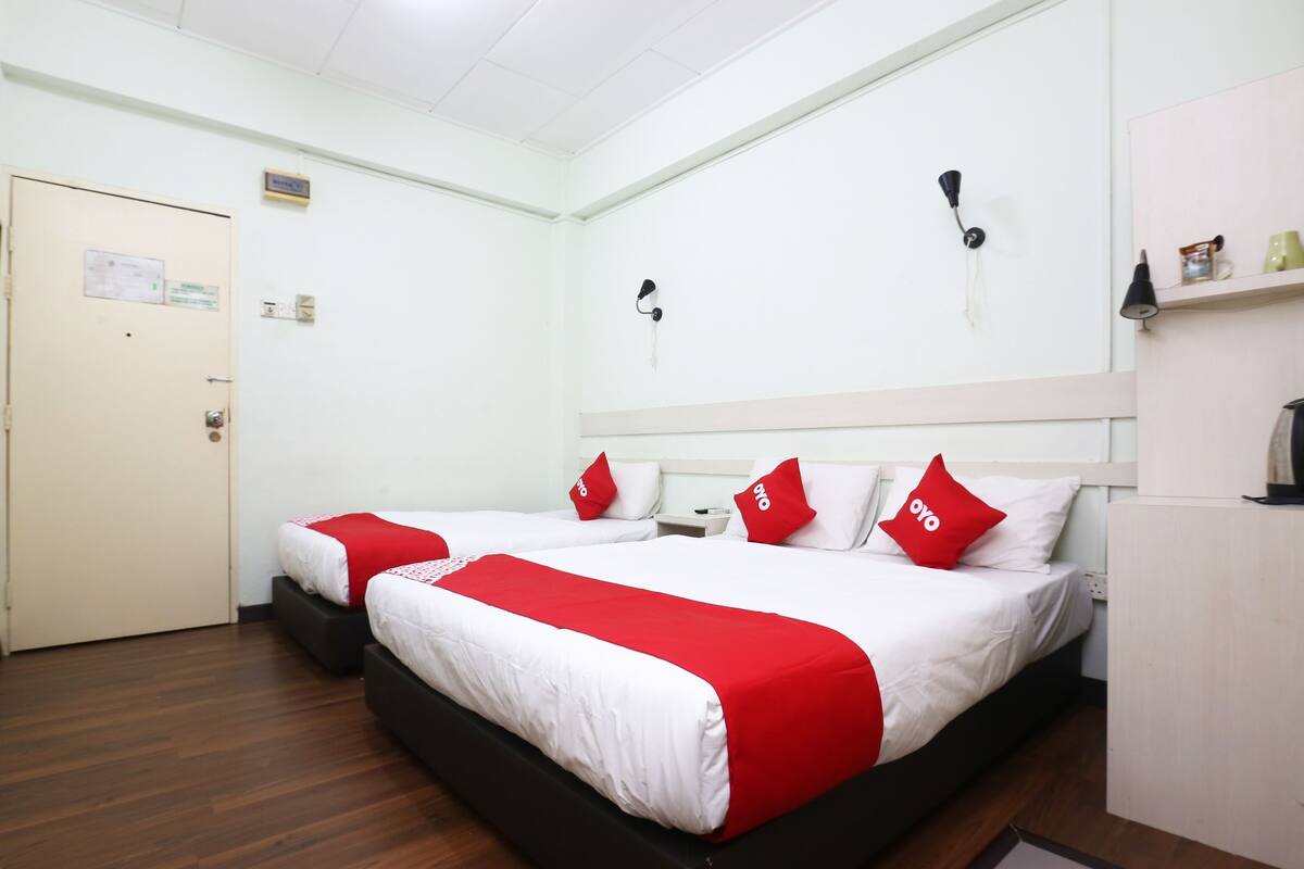 Une chambre d’hôtel avec deux lits, chacun doté de draps blancs et rouges, un plancher en bois, une porte blanche et un petit bureau avec une lampe.