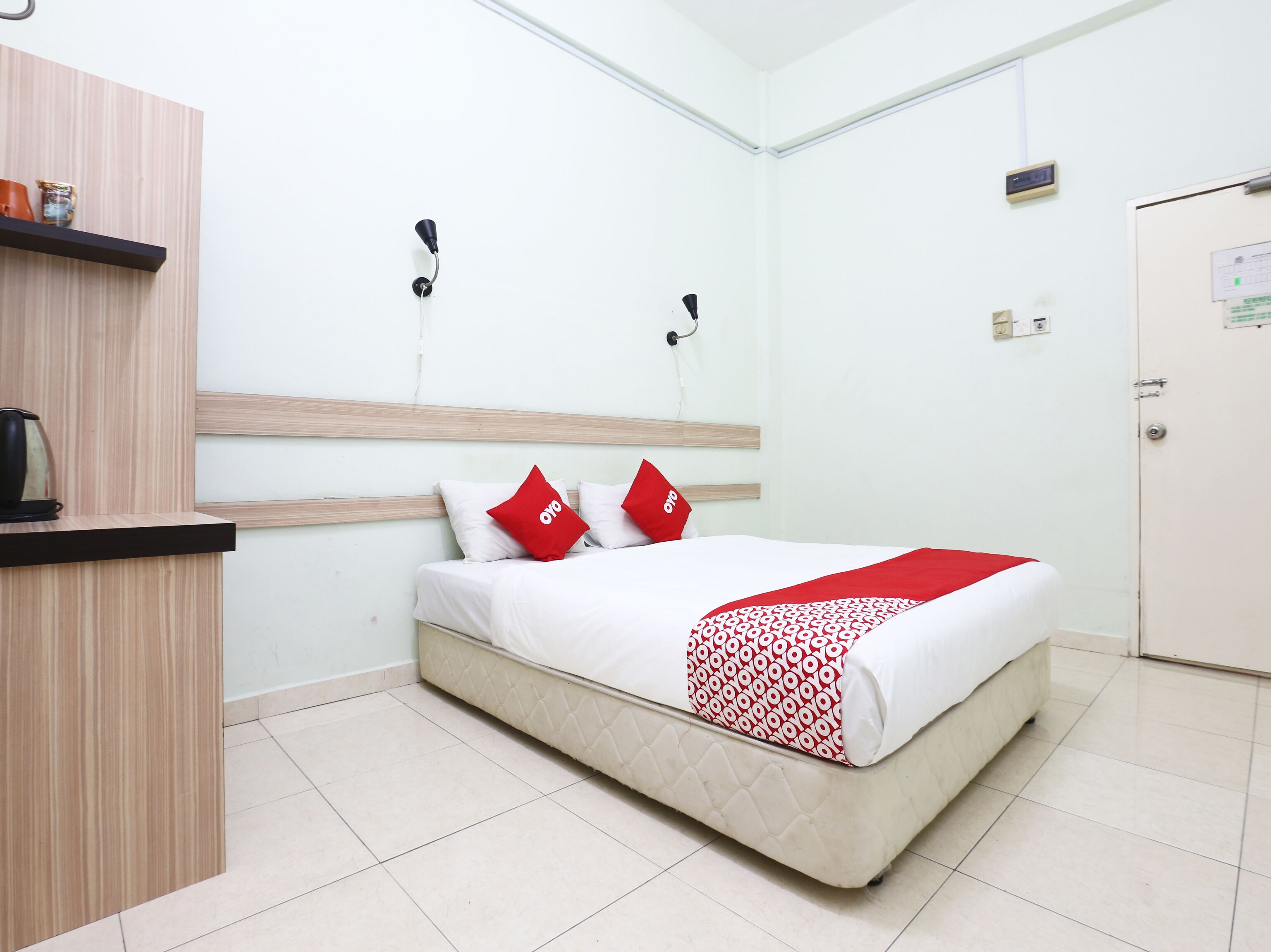 A hotel room with a bed, a wooden headboard, a bedside table, a red and white patterned blanket, and a red pillow.