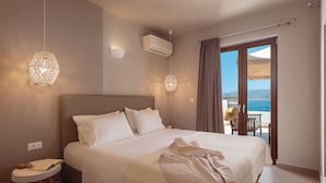 Un dormitorio con una cama grande, una mesita de noche con un libro, aire acondicionado en la pared y vista al mar a través de una puerta de balcón.