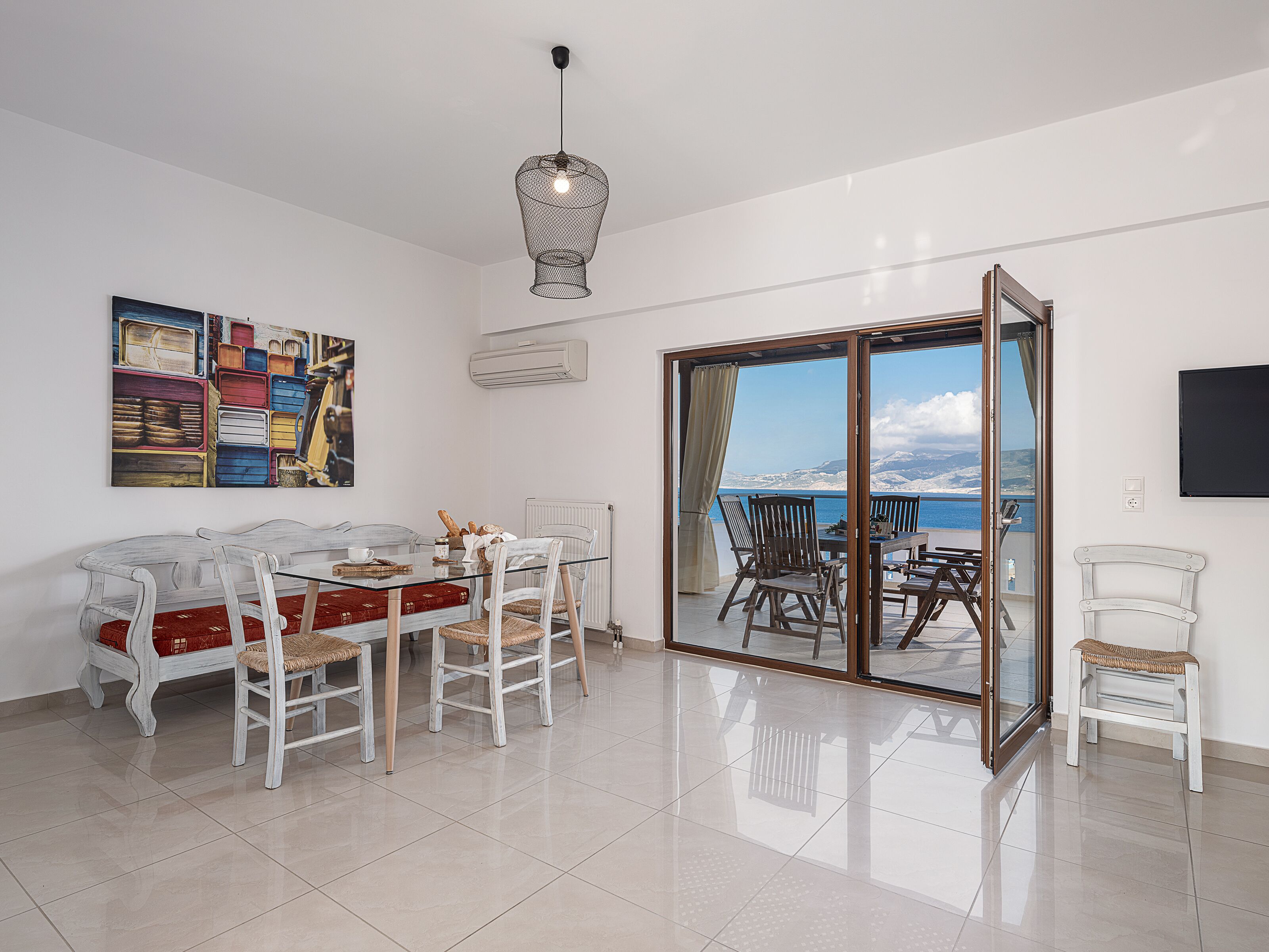 A dining area with a table and chairs, a painting on the wall, and a view of the sea through a glass door.