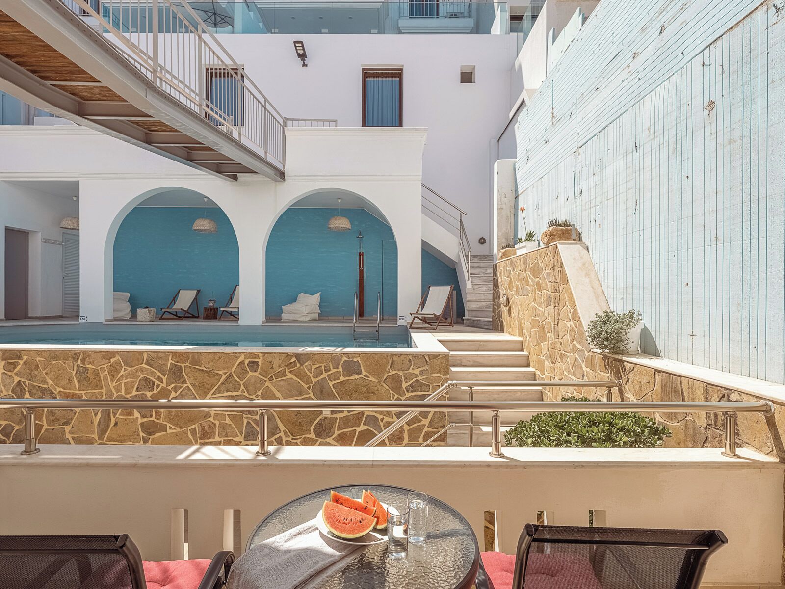Un espace piscine aménagé sur le toit, avec un mur en pierre, une table en verre sur laquelle on a posé une pastèque et des chaises longues.
