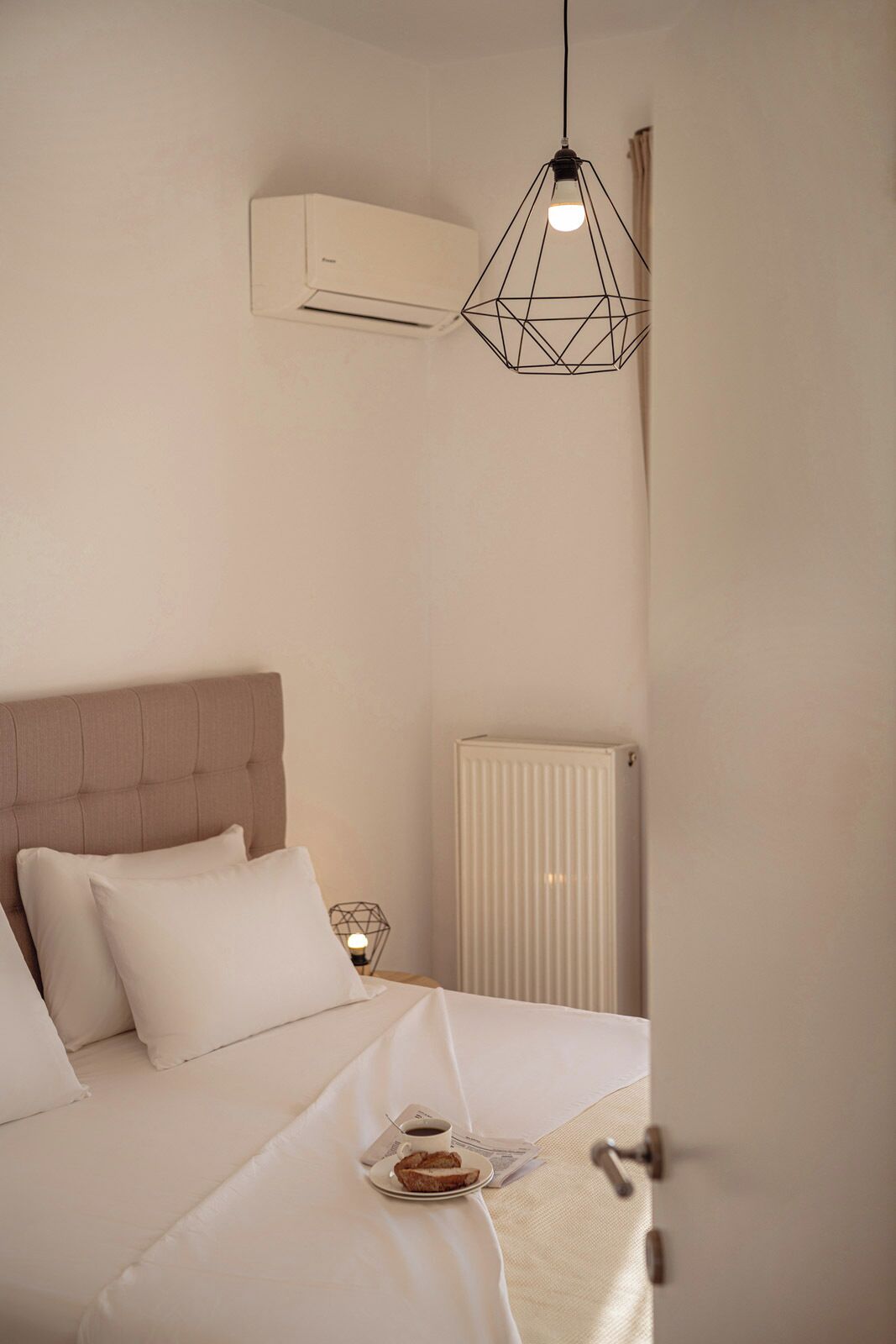 Un lit bien fait, recouvert d’une couette blanche, une table de chevet avec une lampe, et un plateau de petit-déjeuner avec du pain et du café.