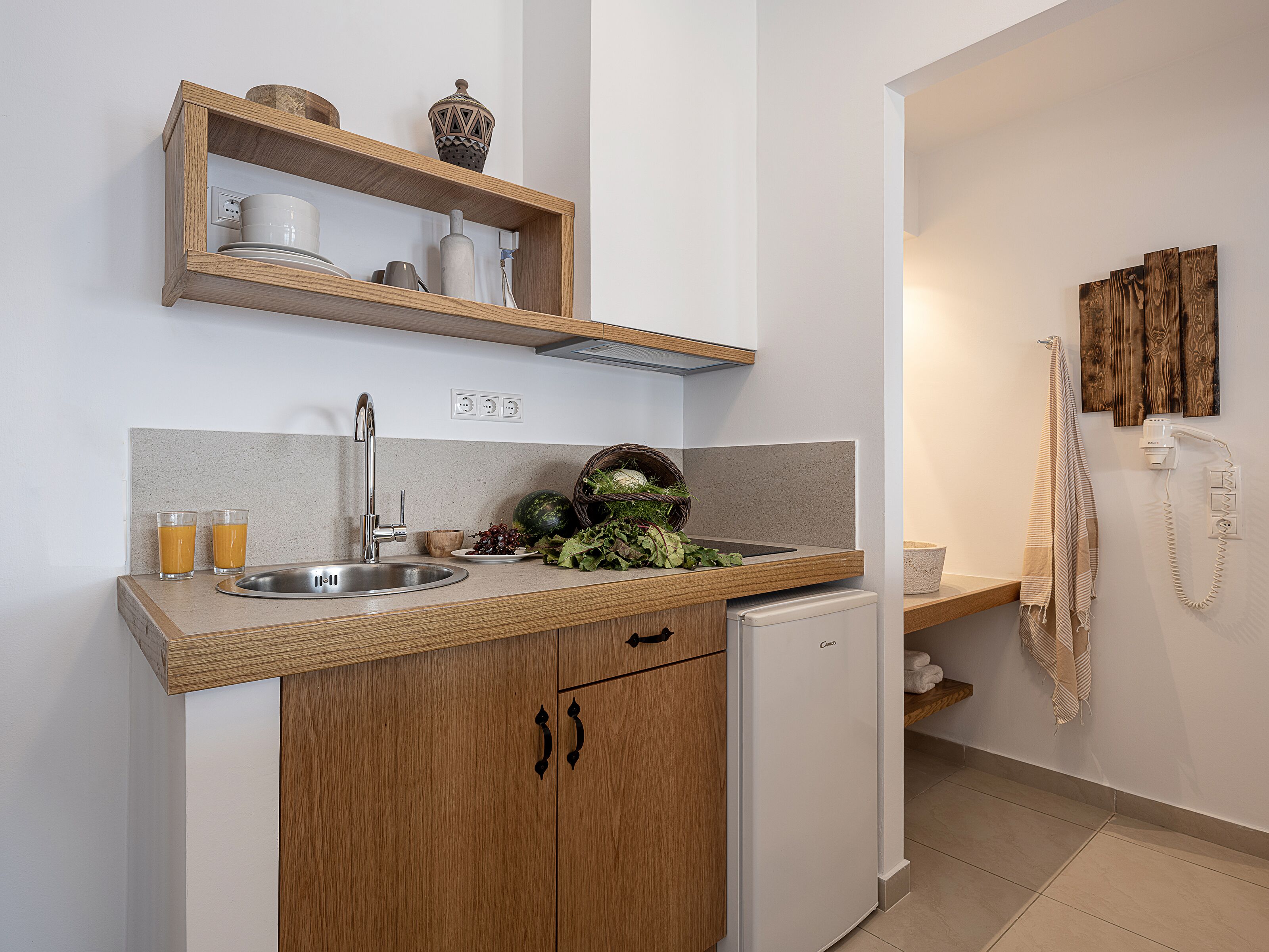 A compact kitchen with a sink, a small refrigerator, wooden cabinets, and a shelf above the counter.