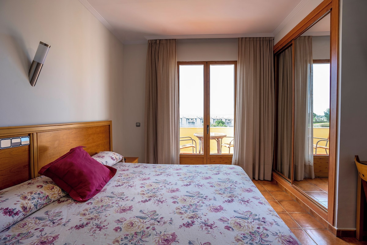 A hotel room with a bed, a wooden headboard, a balcony with a table and chairs, and a sliding glass door.