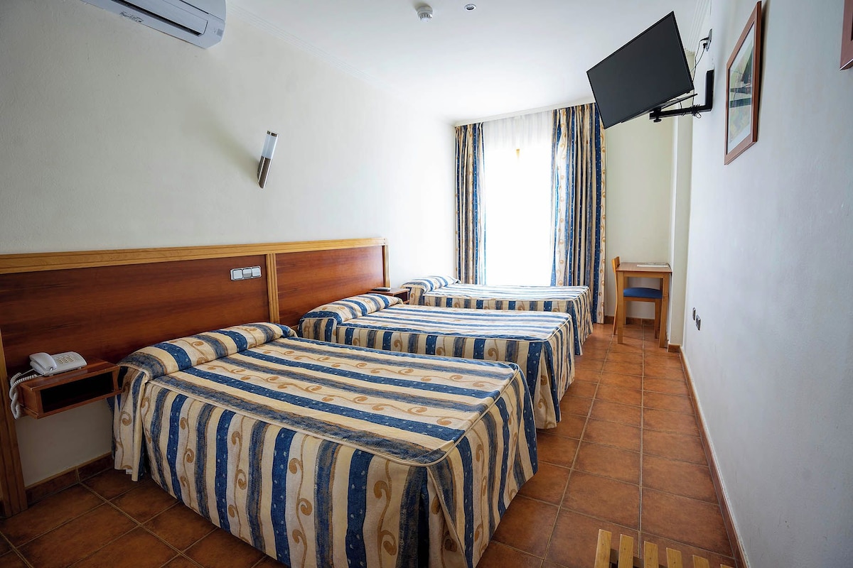 A hotel room with three beds, a TV mounted on the wall, a small table, and a window with curtains.