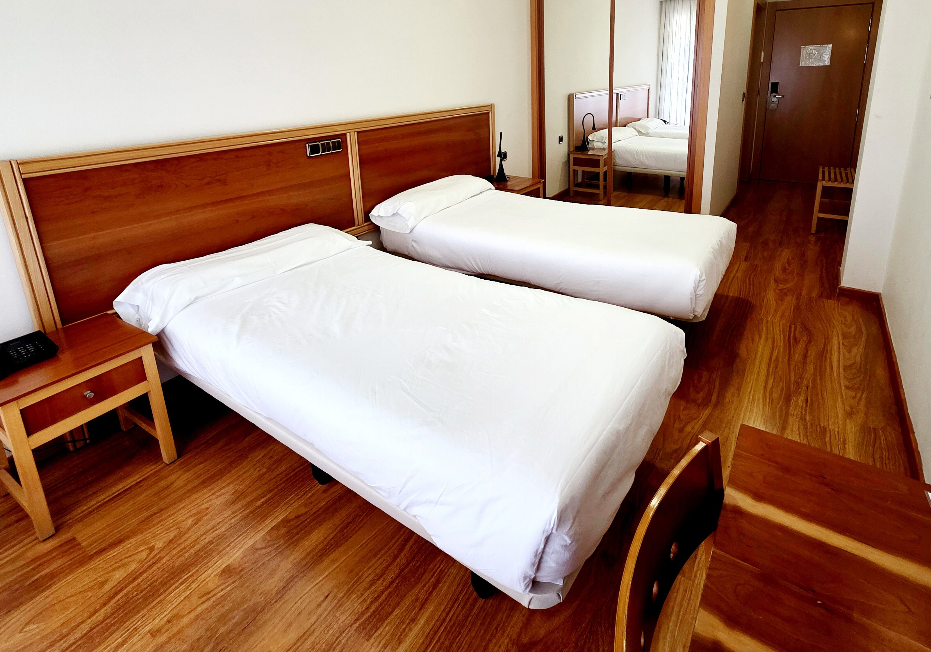 Una habitación de hotel con dos camas individuales, cabeceros y mesita de noche de madera, además de una silla de madera.