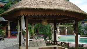 Ein Pavillon mit Strohdach, einem Holztisch und Stühlen, umgeben von einem Pool und Grünflächen.