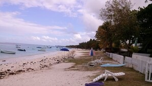 Una playa con varias embarcaciones en el agua, una valla blanca y una fila de sillas reclinables blancas con una sombrilla azul.