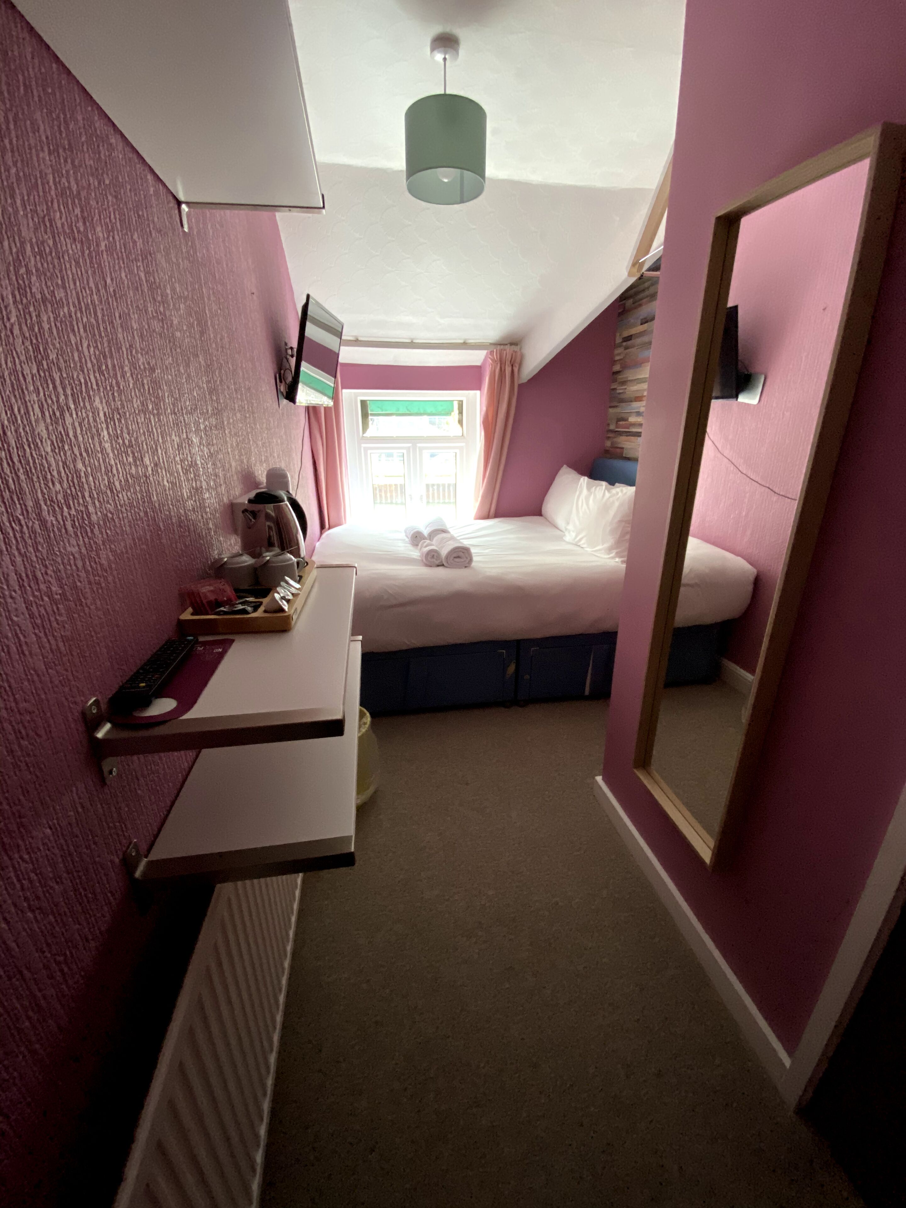 Een kleine slaapkamer met een bed, nachtkastje met lamp, spiegel en raam met gordijnen.