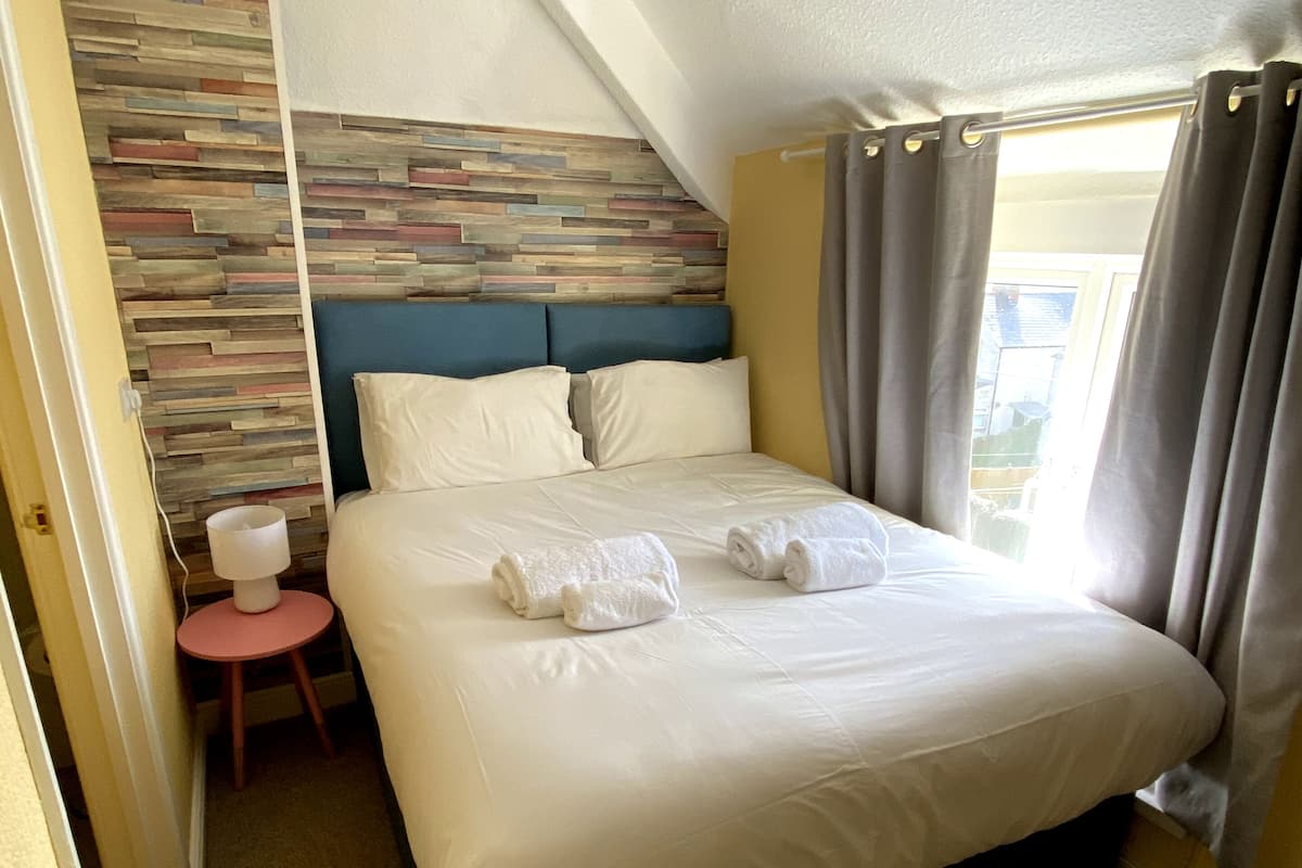 Un lit bien fait, avec du linge de lit blanc et deux serviettes enroulées, une petite table ronde avec une lampe, un mur orné de lambris en bois et une fenêtre avec des rideaux.