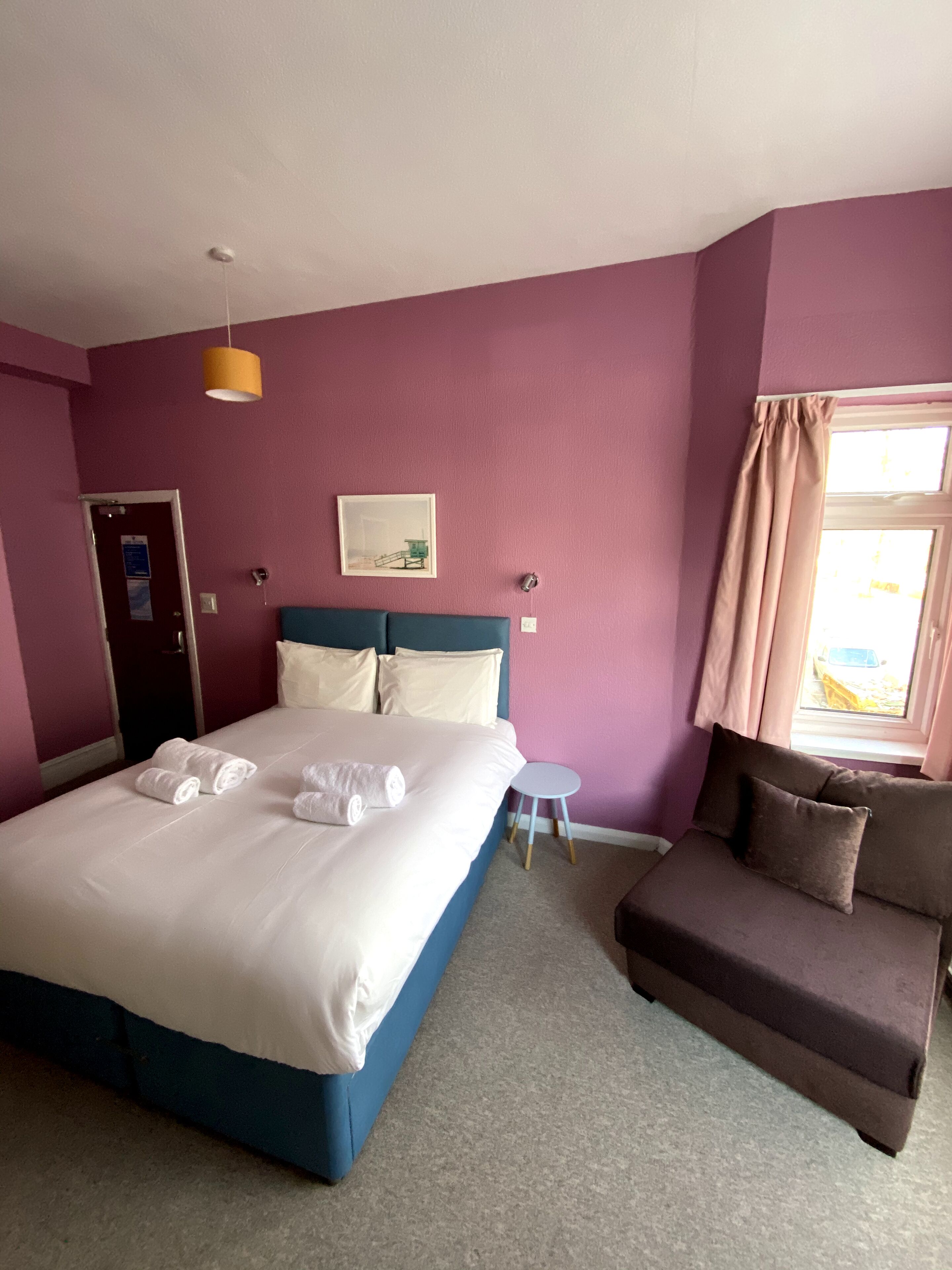 A hotel room with a bed, a small table, a chair, and a window with curtains.