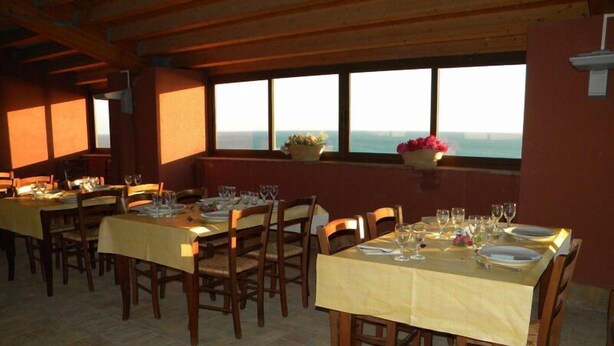 Um restaurante com várias mesas preparadas para refeições, grandes janelas com vista e teto de madeira.