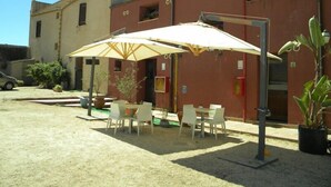 Uma área de estar externa com mesas e cadeiras sob guarda-sóis, um edifício com fachada vermelha e um carro estacionado.