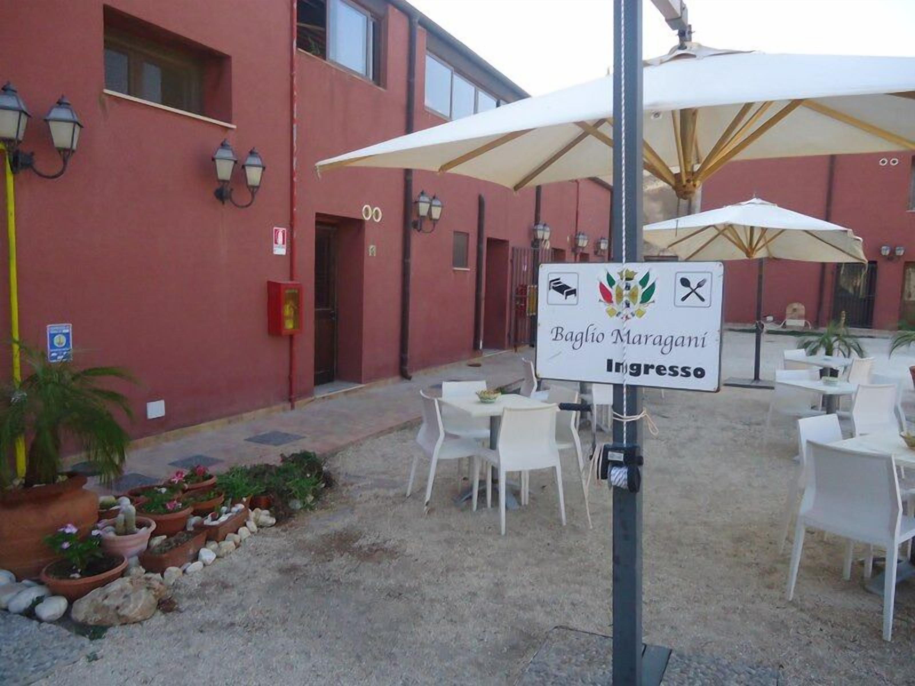 Ein Außenbereich mit weißen Tischen und Stühlen, ein Schild mit der Aufschrift 'Baglìo Maragani ingresso' und ein Gebäude mit roten Wänden und Fenstern.