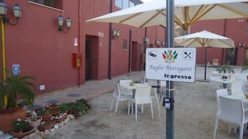 Ein Außenbereich mit weißen Tischen und Stühlen, ein Schild mit der Aufschrift 'Baglìo Maragani ingresso' und ein Gebäude mit roten Wänden und Fenstern.