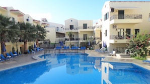 Une piscine aux carreaux bleus, entourée de chaises longues et de parasols, et située à côté d’immeubles à plusieurs étages dotés de balcons.
