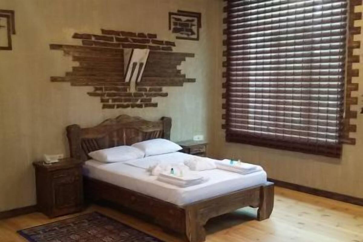 Un lit en bois avec du linge de lit blanc, une table de chevet, une étagère fixée au mur et une fenêtre avec des stores.