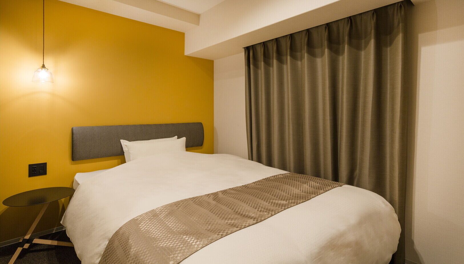 Una habitación de hotel con cama, mesita de noche, lámpara y ventana con cortinas.