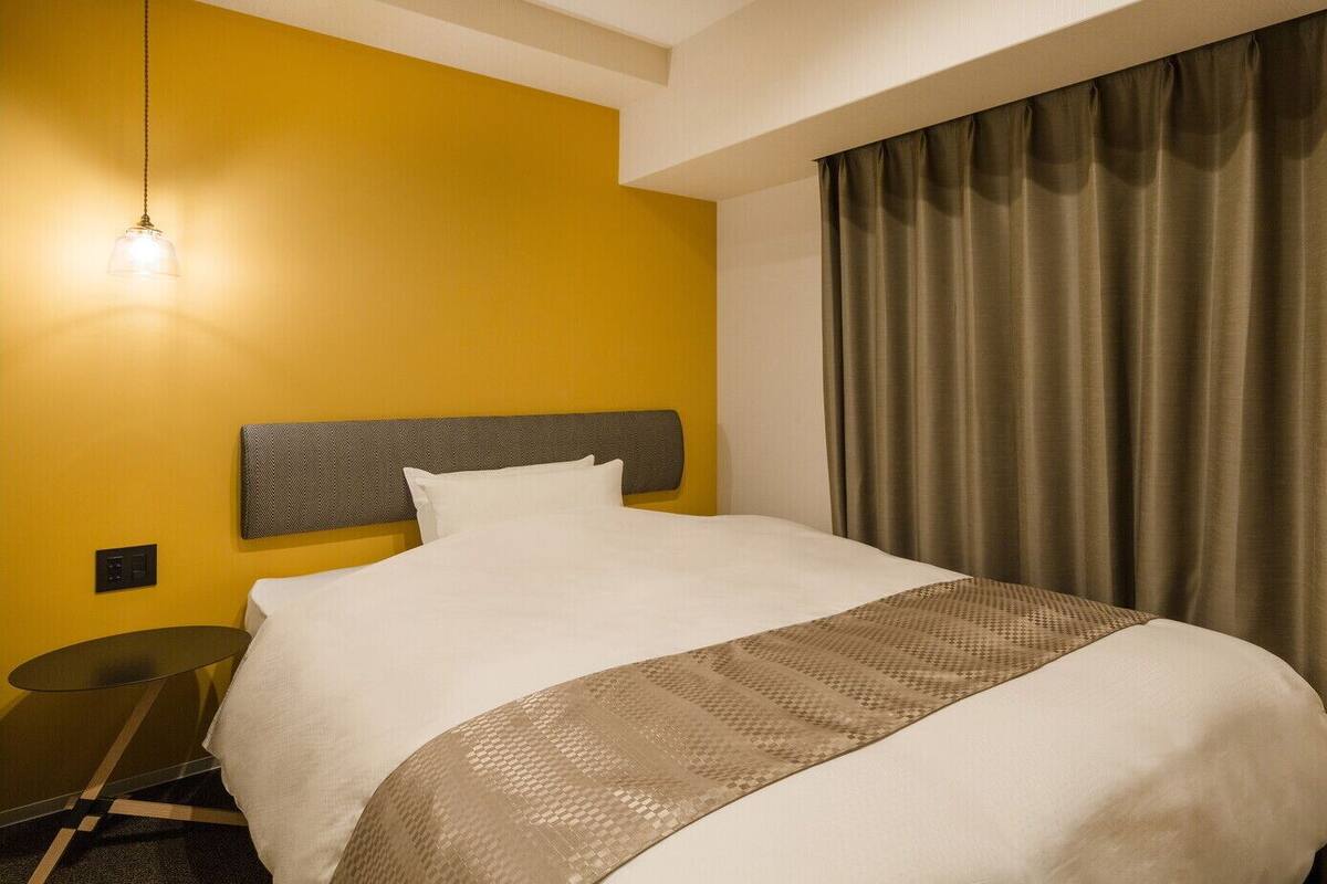 Una habitación de hotel con cama, mesita de noche, lámpara y ventana con cortinas.