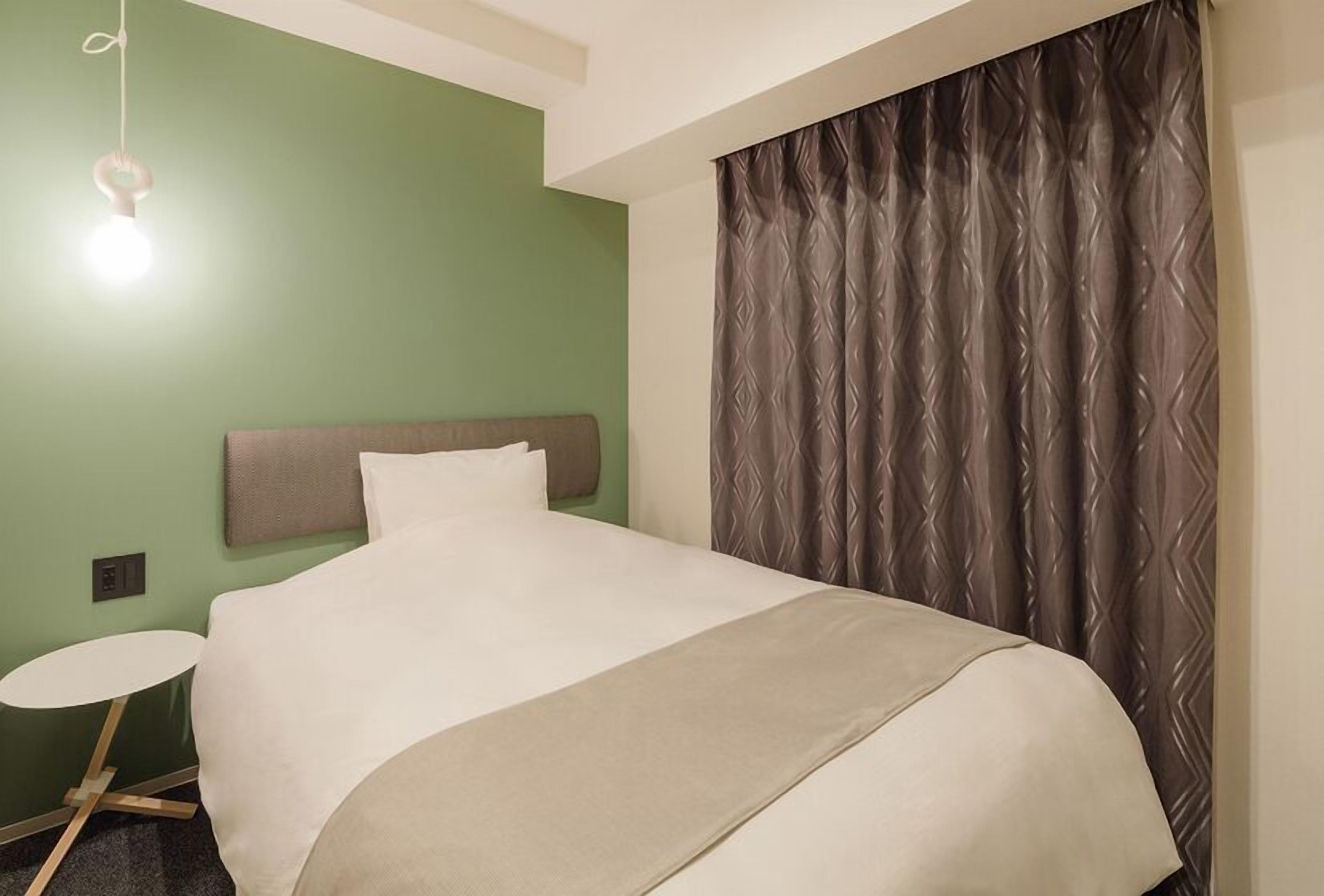 Habitación de hotel con una cama, una mesa redonda, un aplique de pared y una cortina estampada.