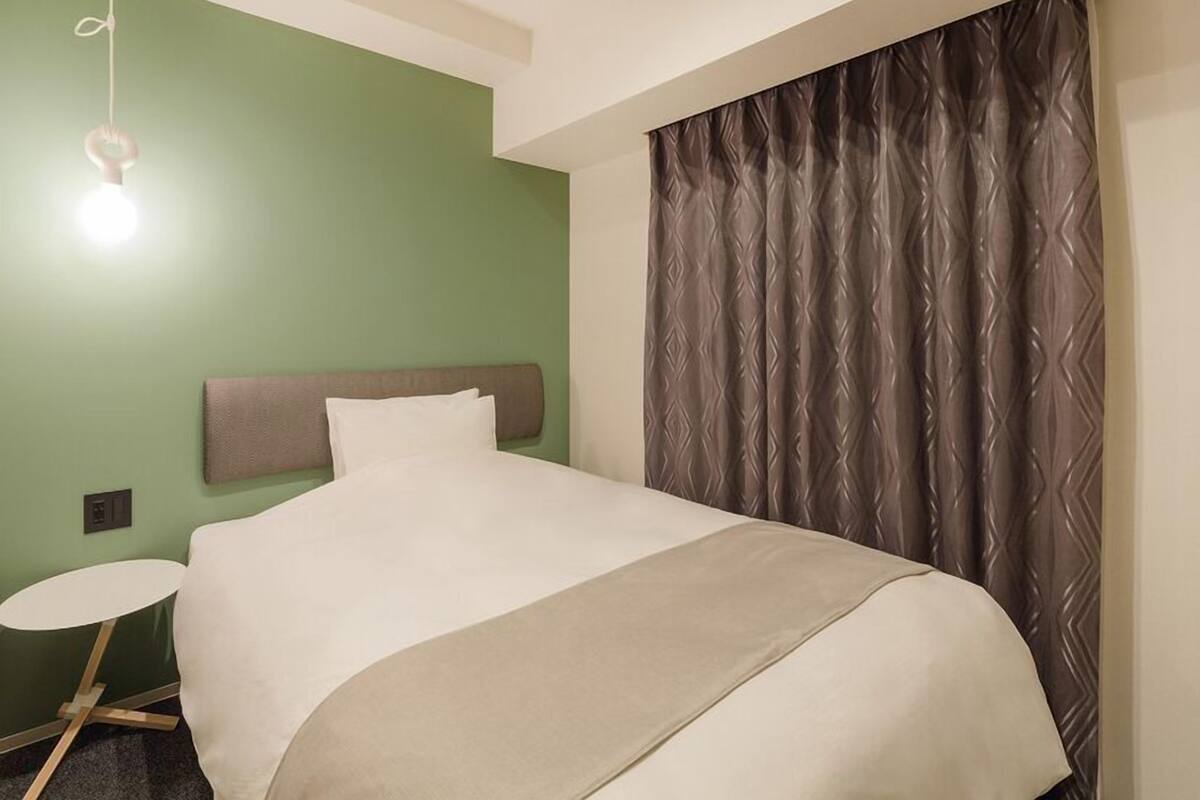 Une chambre d’hôtel avec un lit, une table ronde, un luminaire fixé au mur et un rideau à motifs.