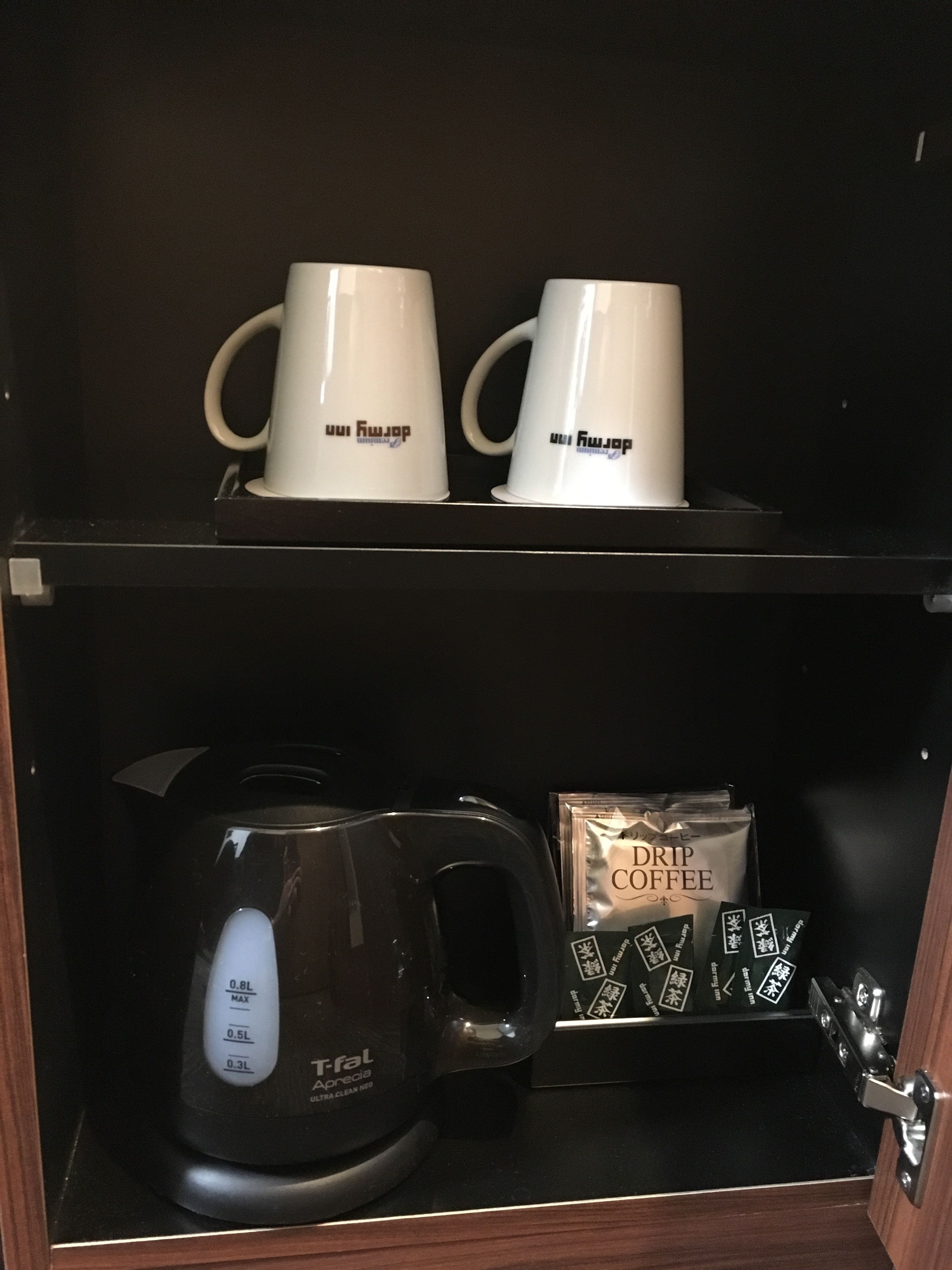 A coffee maker, two white mugs, and drip coffee packets in a cabinet.