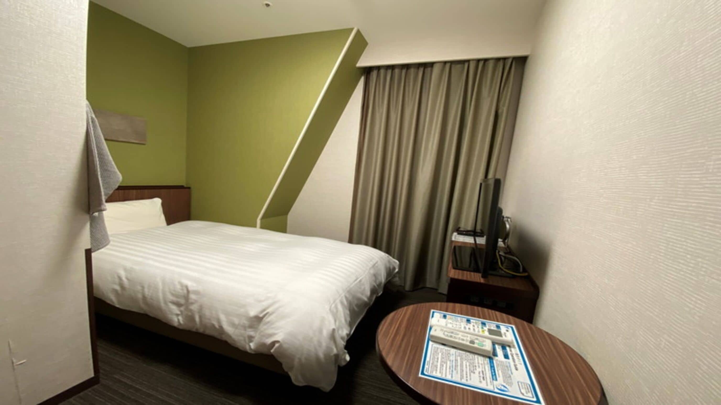 Una habitación de hotel con una cama, una mesita de noche con un control remoto, una pequeña mesa redonda con un folleto y una ventana con cortinas.