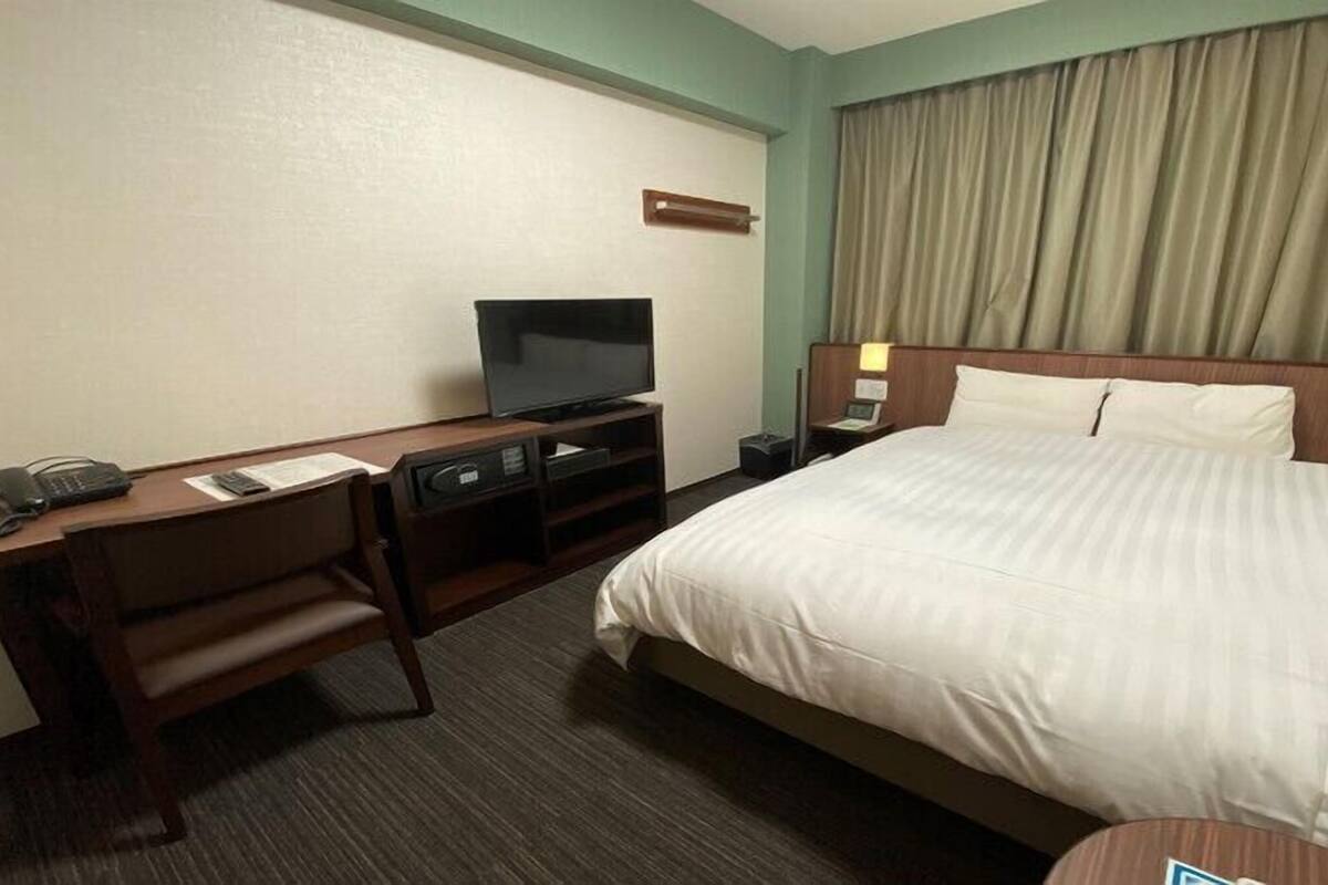 A hotel room with a bed, a desk with a chair, a television, and a nightstand with a lamp.