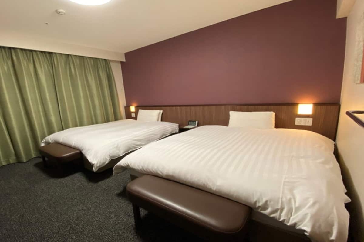 A hotel room with two beds, a bench, a desk, and a green curtain.