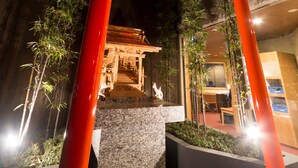 Un torii japonés tradicional con una placa dorada, rodeado de plantas de bambú y un sendero de piedra que conduce a la entrada de un edificio.