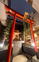Un torii japonés tradicional con una placa dorada, rodeado de plantas de bambú y un sendero de piedra que conduce a la entrada de un edificio.