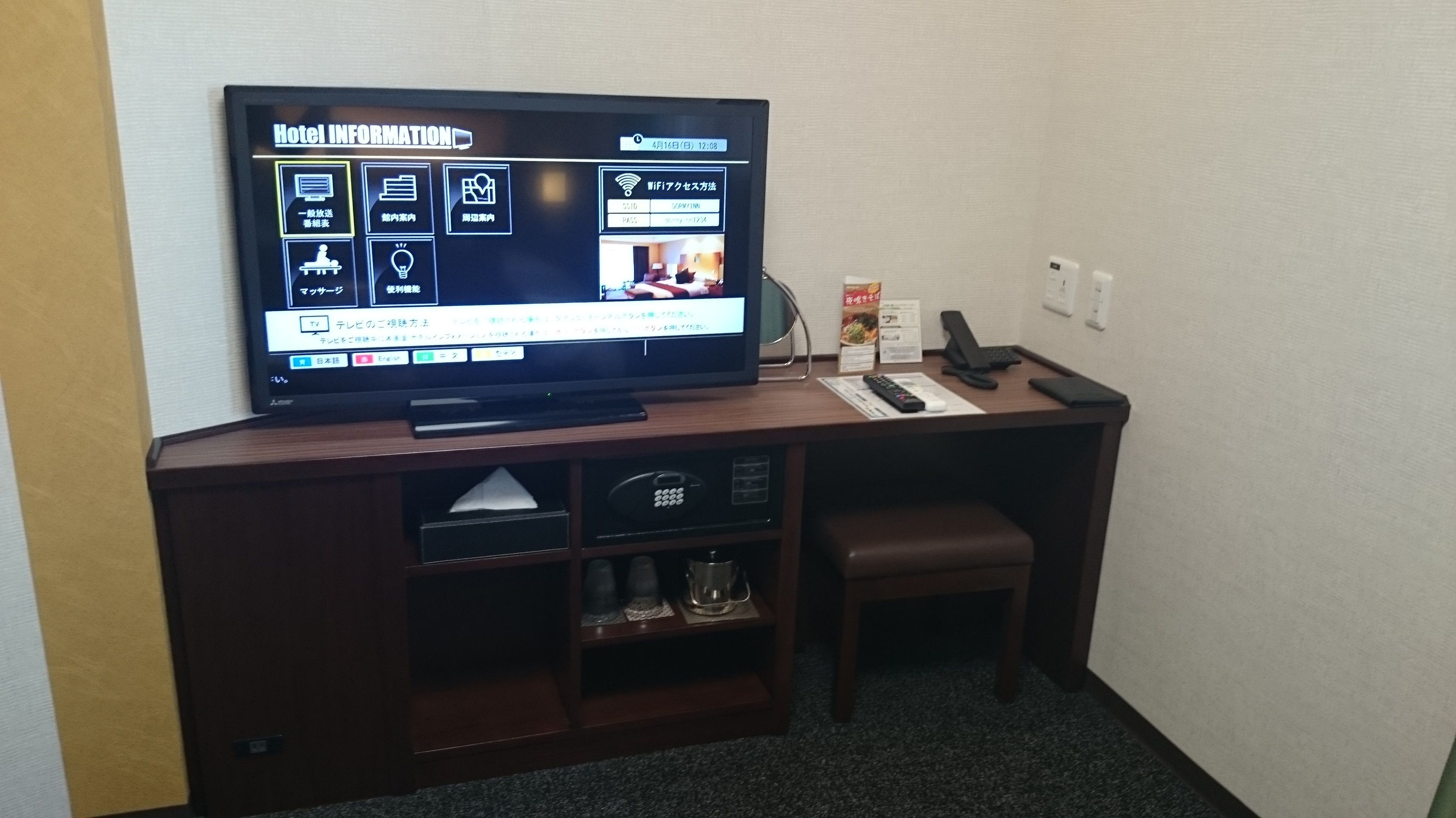 A hotel room with a flat-screen TV displaying hotel information, a desk with a keyboard and mouse, a telephone, and a small seating area.