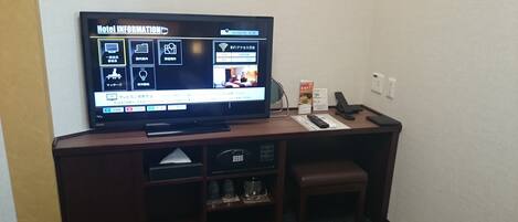 A hotel room with a flat-screen TV displaying hotel information, a desk with a keyboard and mouse, a telephone, and a small seating area.