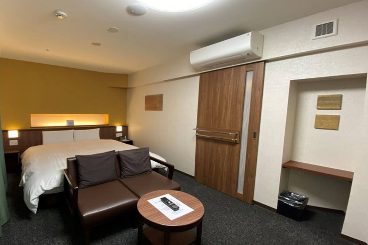 A hotel room with a bed, a sofa, a small table with a remote control, an air conditioning unit, and a trash bin.