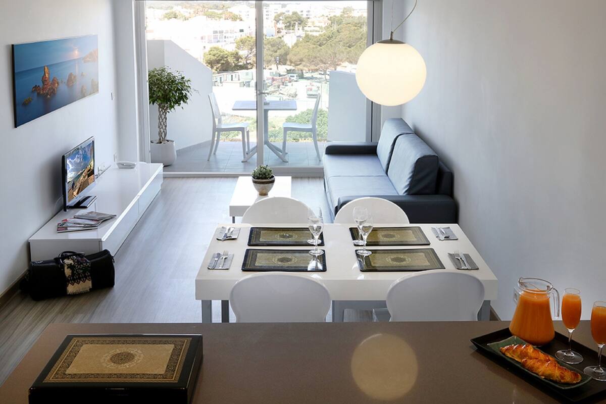 Uma sala de jantar moderna com uma mesa branca posta para dois, um sofá e vista para a cidade através de grandes janelas.