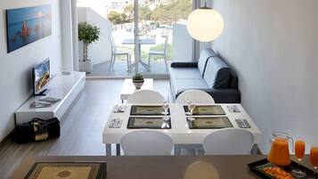 A modern dining area with a white table set for two, a sofa, and a view of the city through large windows.