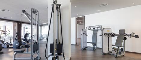 A modern gym with various exercise machines, a wooden floor, and large windows allowing natural light.