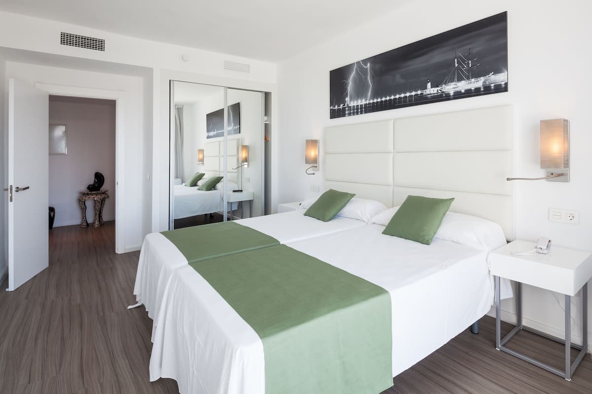 Quarto de hotel com uma cama grande, mesa de cabeceira com telefone, espelho e quadro de um navio na parede.