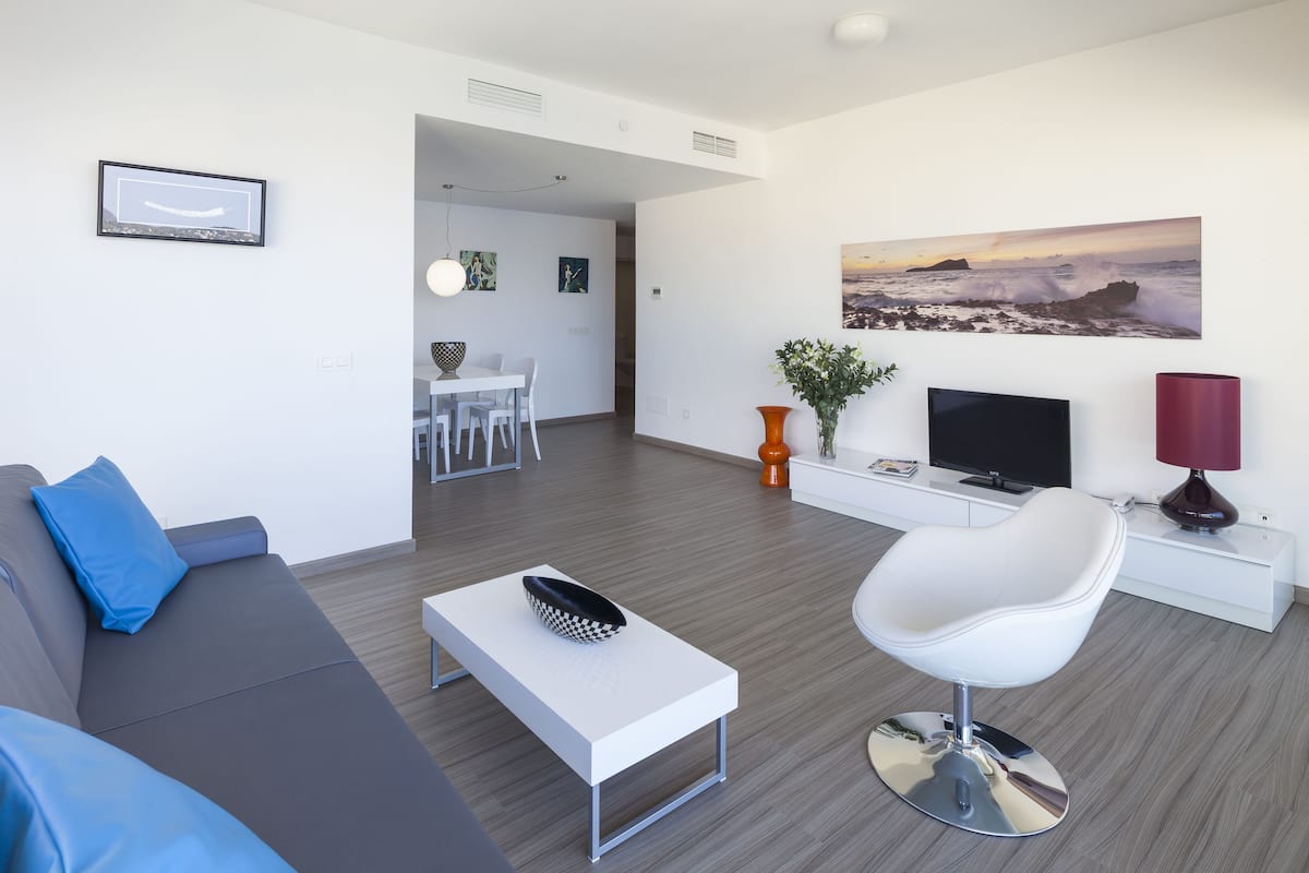 Uma sala de estar moderna com um sofá cinza, uma mesa de centro branca, uma cadeira branca, uma TV e um quadro com uma paisagem marítima.
