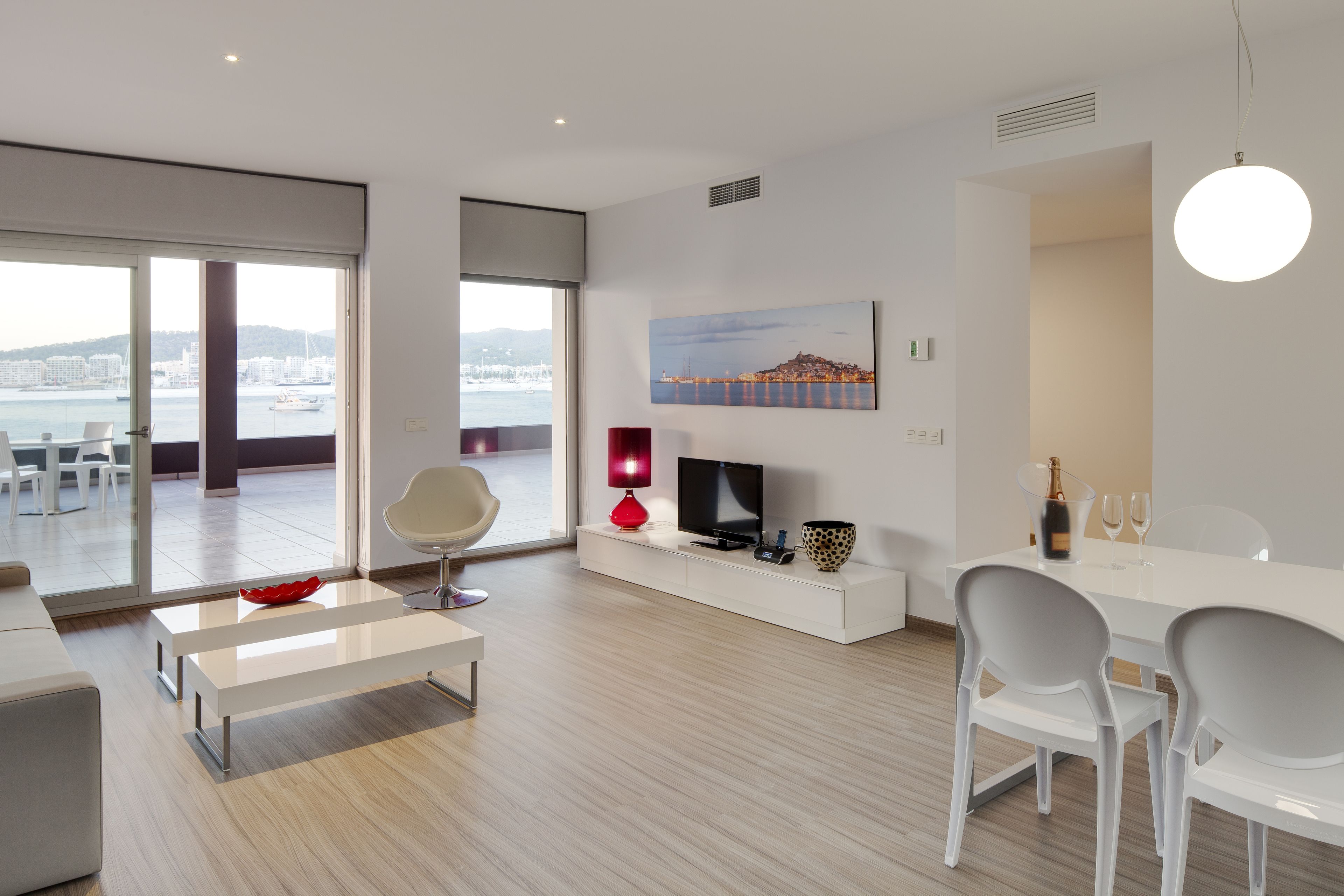 Una sala de estar moderna con un ventanal grande, un televisor de pantalla plana, una lámpara roja y un comedor con sillas blancas y una mesa.