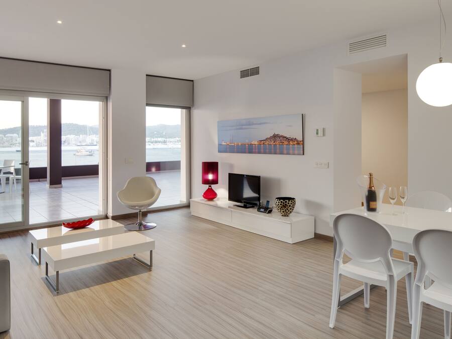 A modern living room with a large window, a flat-screen TV, a red lamp, and a dining area with white chairs and a table.