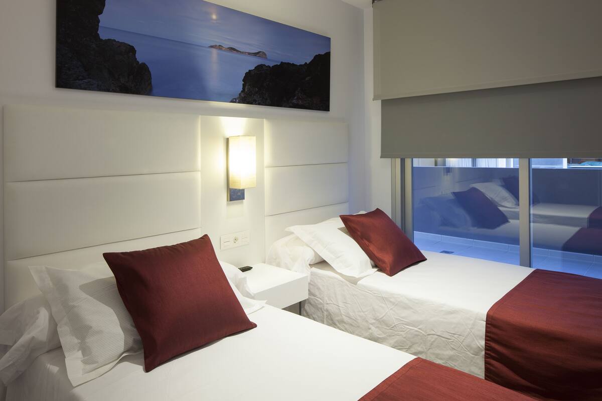 Quarto de hotel com duas camas, uma mesa de cabeceira, uma luminária na parede e uma janela grande com vista para o mar.