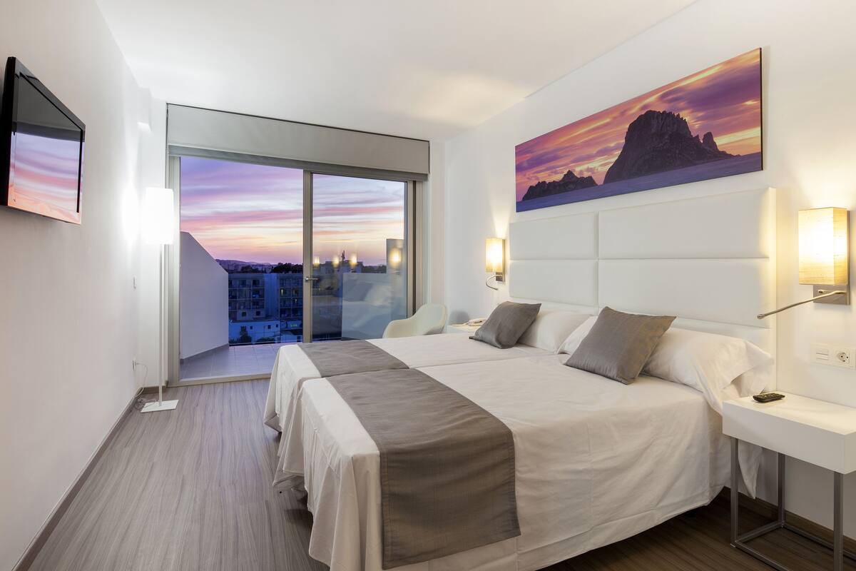 Quarto de hotel com duas camas, uma janela grande com vista para a cidade, um quadro na parede e uma escrivaninha com uma luminária.