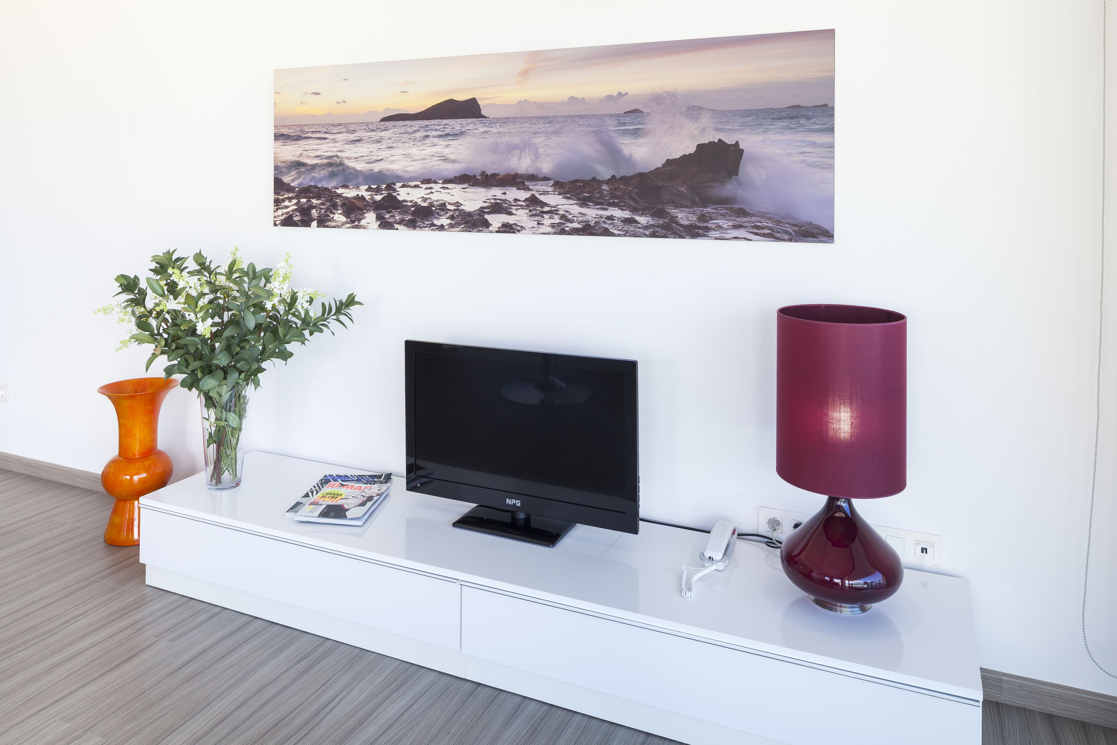 Una sala de estar moderna con un televisor de pantalla plana, una planta decorativa, una revista y una lámpara roja sobre una consola blanca.