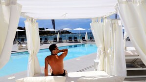 A poolside area with a person relaxing under a canopy, lounge chairs, and umbrellas in the background. - Axel Beach Ibiza - Adults Only (Sant Josep de sa Talaia)