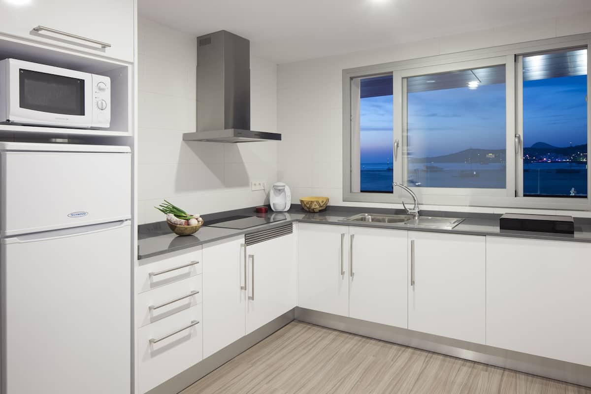 Uma cozinha moderna com armários brancos, micro-ondas, fogão e janela com vista para o mar.
