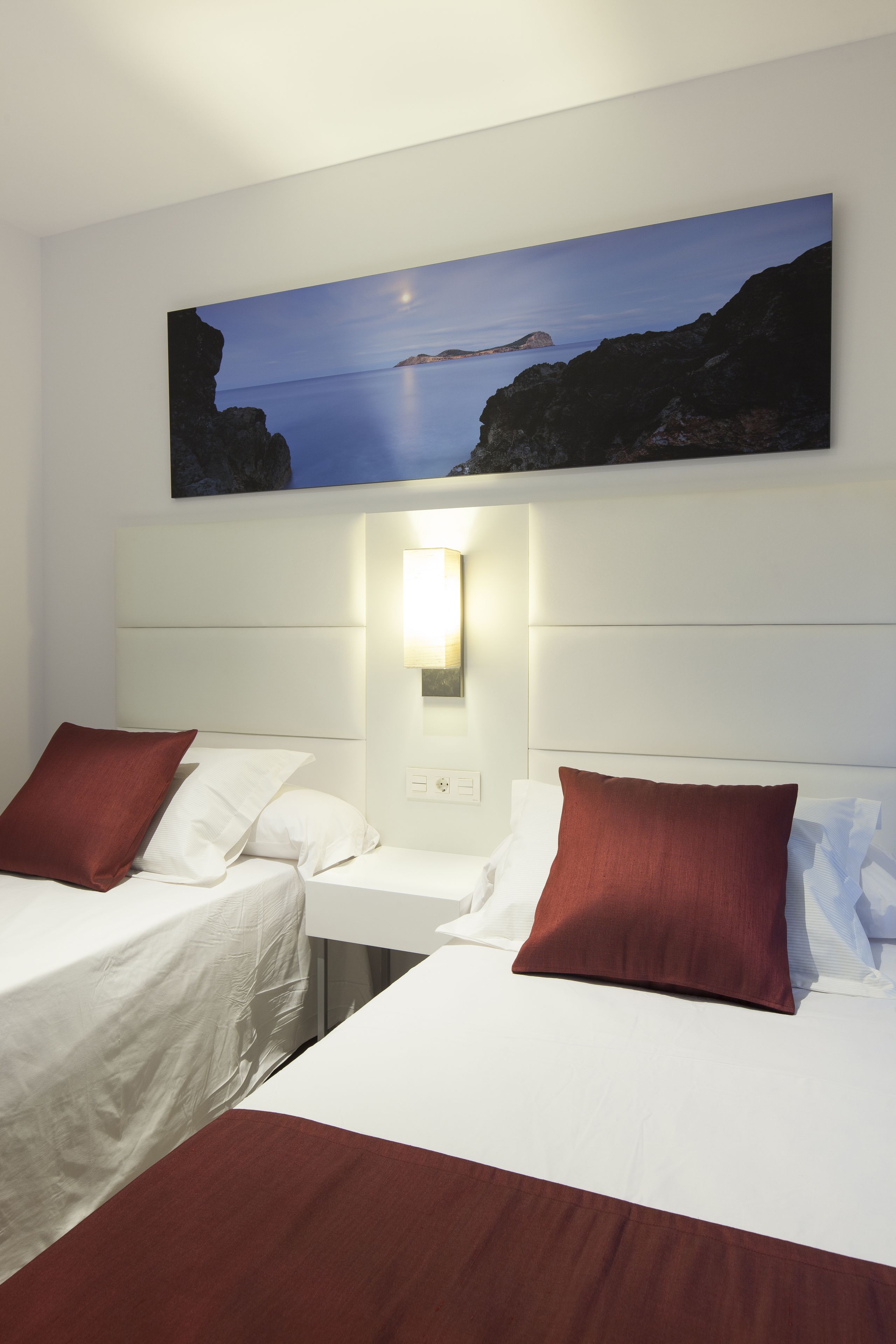 A hotel room with two beds, a nightstand, a wall-mounted lamp, and a painting of a coastal scene.