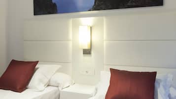 A hotel room with two beds, a nightstand, a wall-mounted lamp, and a painting of a coastal scene.