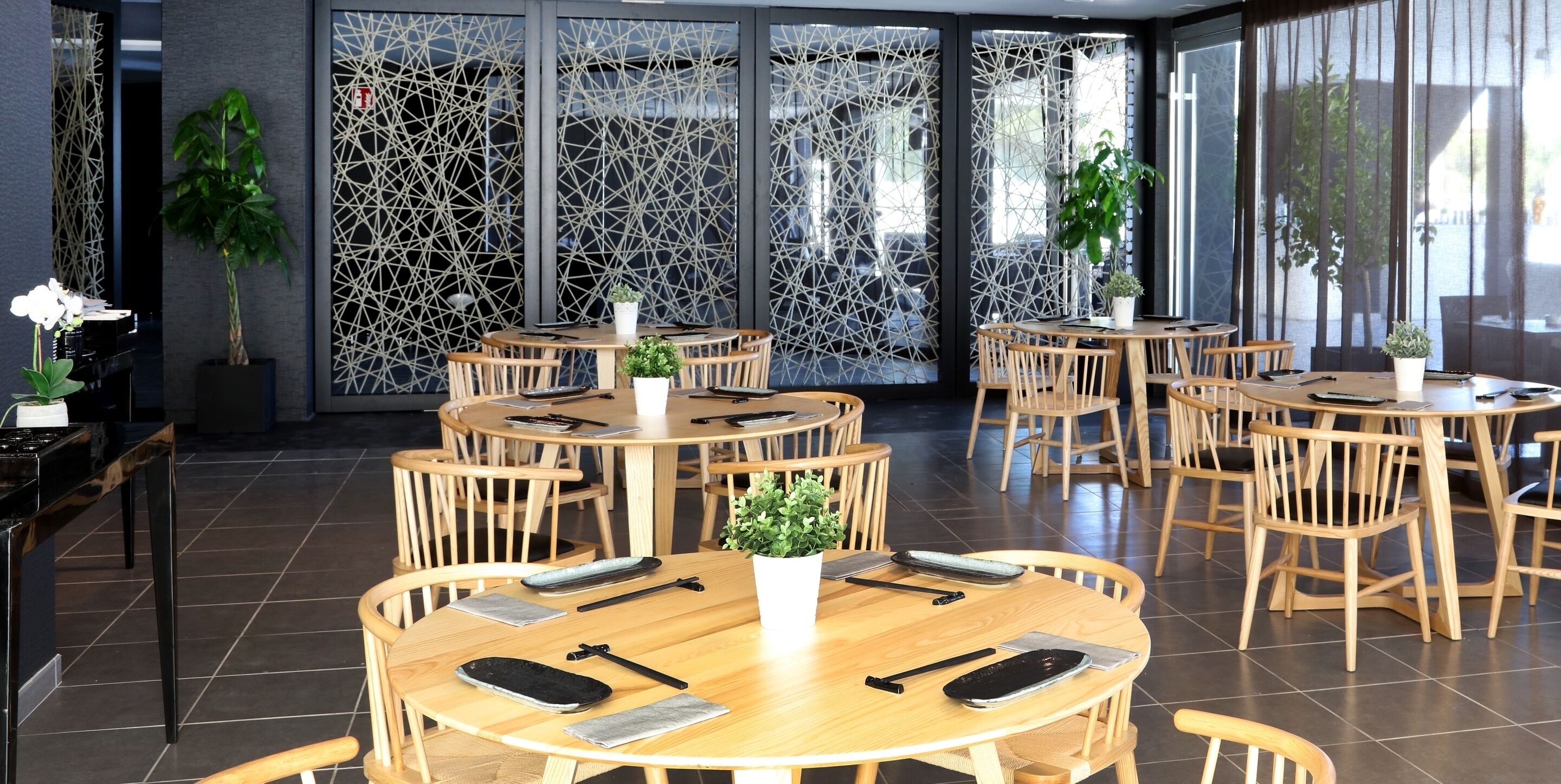 A modern dining area with round wooden tables and chairs, potted plants, and a decorative screen with a geometric pattern.