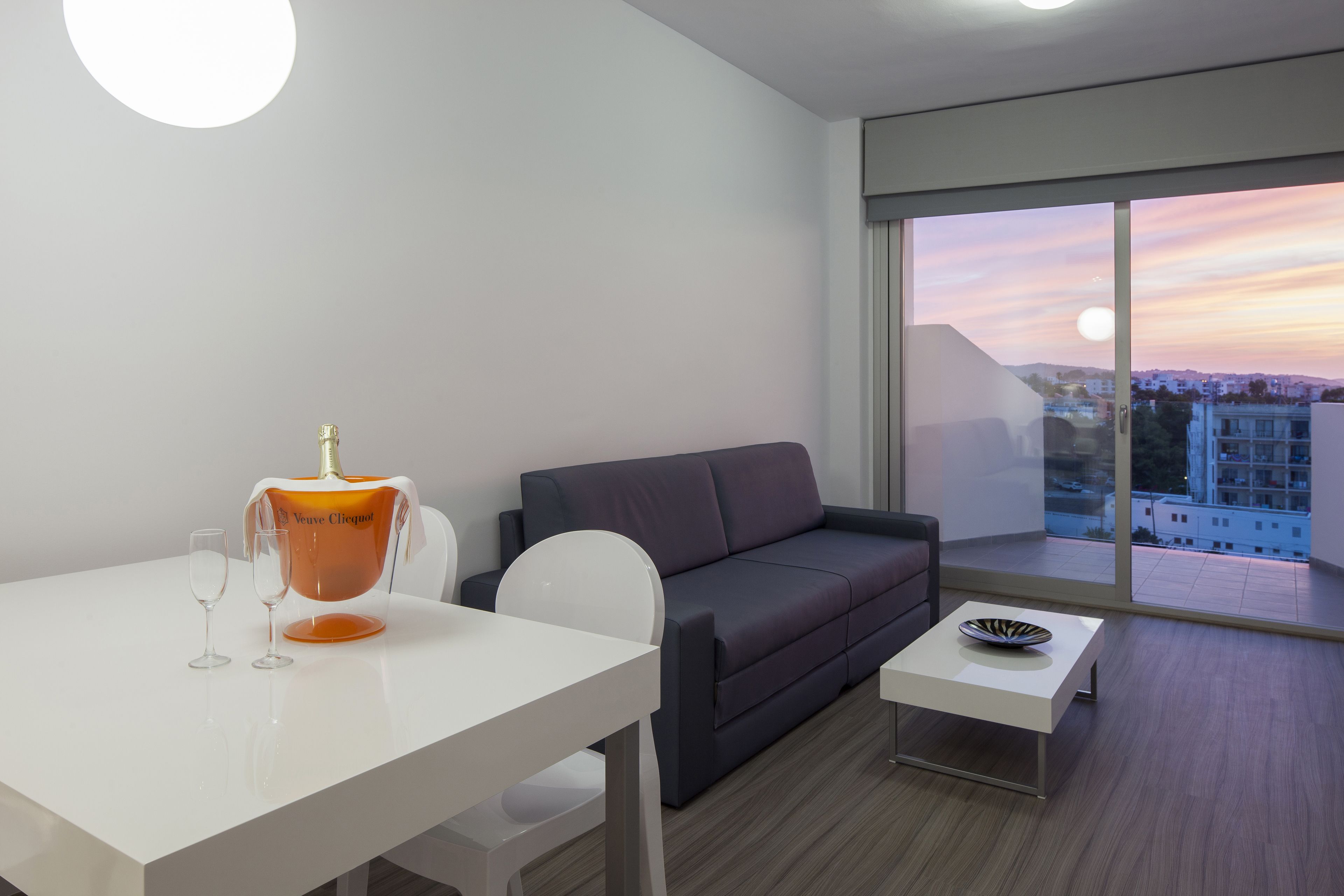 A modern living room with a white table, two wine glasses, a bottle of Veuve Clicquot, a dark grey sofa, and a small white side table with a decorative bowl.